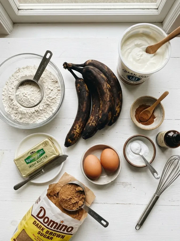 Overhead flat lay, four heavily blackened overripe bananas centred, open tub of thick white Greek yogurt, two eggs, glass bowl of all-purpose flour, stick of butter, bag of brown sugar, small bowls of cinnamon and baking soda, clean white background, soft even natural light