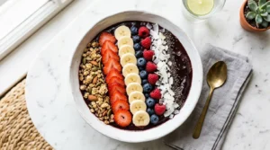 Overhead angle, wide white bowl filled with deep purple-black thick acai smoothie base, beautifully arranged rows of granola, fresh sliced strawberries, banana coins, shredded coconut, and chia seeds, white marble surface, bright natural window light