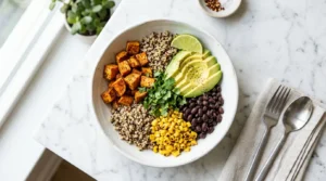 Overhead angle, wide white bowl filled with golden roasted sweet potato cubes, black beans, charred corn, sliced avocado, and fluffy quinoa arranged in neat sections, fresh lime wedge and cilantro scattered on top, white marble surface, bright natural window light